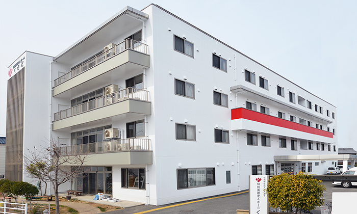 白壁4階建ての介護施設。バルコニー付き、赤い看板が目印。駐車場完備。